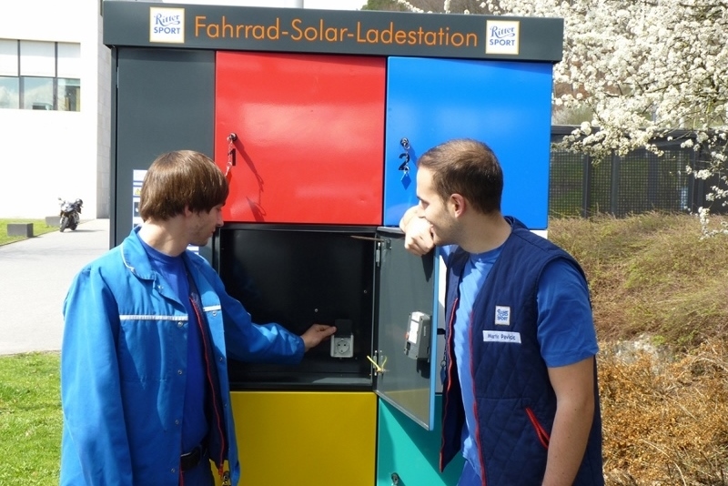 Zwei Männer stehen vor einer bunten Fahrrad-Solar-Ladestation mit Fächern in Blau, Rot, Gelb und Grün. 