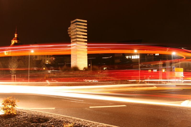 Straße bei Nacht mit beleuchteter Skulptur und Lichtspuren. 